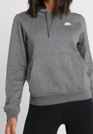 Kobieta ubrana w szarą bluzę Nike z kapturem, kieszenią z przodu i sznureczkami, w połączeniu z czarnymi legginsami.