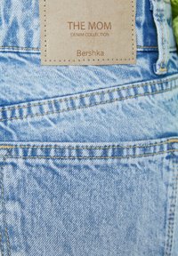 Světle modré džíny z denimu s výraznou vybledlou texturou, zlatým prošíváním a hnědou štítkem s nápisem "THE MOM DENIM COLLECTION" od Bershky.