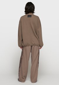 Brauner, übergroßer Sweatshirt mit strukturierter Oberfläche, versehen mit einem kleinen schwarzen Etikett auf der Rückseite. Kombiniert mit weiten Hosen in einer passenden Farbe.