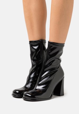 Schwarze Stiefeletten aus Lackleder mit einem Blockabsatz, vorne mit Reißverschluss und geraffter Textur am Schaft. Glatte, glänzende Oberfläche.