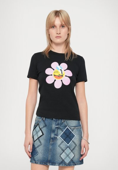 Γυναίκα φορώντας ένα μαύρο t-shirt με σχέδιο ροζ λουλούδι και χαμογελαστό emoji, συνδυασμένο με μπλε τζιν φούστα με διαμαντένια πατσαβούρα.
