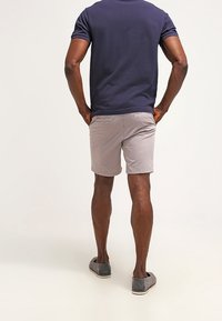 Homme debout les mains dans les poches arrière, portant un polo bleu marine, un short gris clair et des chaussures grises sans lacets sur un fond uni.
