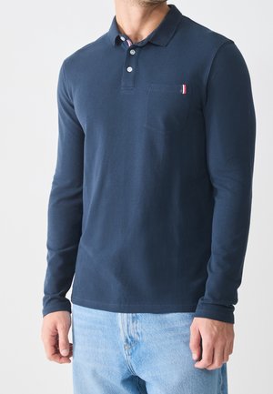 Man in een marineblauwe polo met lange mouwen en een klein zakdetail, gedragen met lichtblauwe denim jeans, tegen een effen achtergrond.