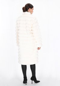 Witte nepbontjas met een geribbeld ontwerp, hoge kraag en een middellange snit. Ggedragen over zwarte leggings en stijlvolle zwarte hakken.