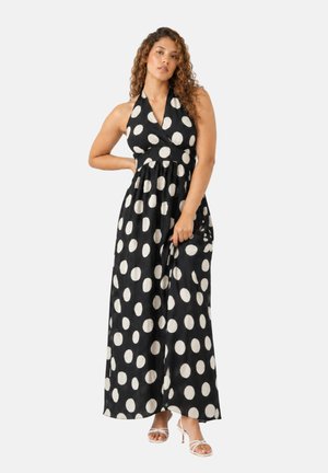 Frau mit lockigen Haaren, die ein schwarzes Maxikleid mit großen weißen Punkten und weißen Riemchen-High Heels trägt.