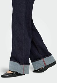 Temno modre široke hlače iz trapera z kontrastnim svetlo modrim robom in rdečim šivom, kombinirane s črnimi ravnimi čevlji z dodatki zakovic.