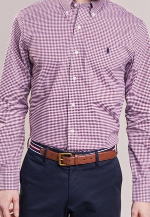 Mann trägt ein lila-weiß kariertes Hemd, in navyblaue Hose gesteckt, mit braunem Ledergürtel und gestreiftem Gürtelschlaufen-Detail.