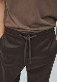 Brązowe bawełniane spodnie o prostym kroju, z regulowanym sznurkiem w pasie, zapinane na guziki, z bocznymi kieszeniami. Gładka faktura.