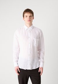Camisa de lino blanca de manga larga con ligera textura, bolsillo en el pecho y dobladillo redondeado, combinada con pantalones oscuros.