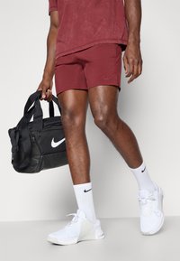 Zwarte Nike sporttas, bordeauxrode shorts, witte sport schoenen en bijpassende sokken. Materiaal lijkt glad; ontwerp is sportief en functioneel.