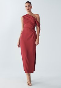 Viens pleca sarkana kleita no mīksta auduma ar savāktu sānu detaļu un sānu šķēlumu, papildināta ar zelta siksnu sandales.