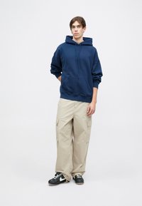 Granatowy sweatshirt z kapturem i kieszenią typu kangur, beżowe spodnie cargo z kieszeniami bocznymi oraz czarne trampki z białymi akcentami.