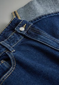 Tumši zili džinsi ar kontrastējošu gaiši zilu jostu, sudraba pogu un redzamu dzeltenu šuvju dekorējumu gar šuvēm un kabatām.