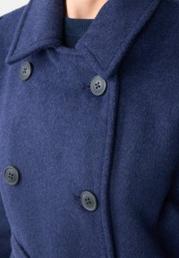 Dubbelzijdige marineblauwe wollen jas met grote zwarte knopen, gestructureerde kraag en geribbelde gebreide halslijn. Zachte textuur en op maat gemaakte pasvorm.