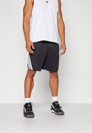 Männliche Person trägt schwarze Shorts mit weißen Streifen, weißes ärmelloses Sportshirt, weiße Socken und schwarze Sportschuhe und steht vor einem einfarbigen Hintergrund.