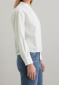 Valkoinen puuvillapaita, jossa on pystykaulus, kaksi nappia, pitkät hihat ja hieman lyhyt pituus. Yhdistettynä sinisiin farkkuihin.