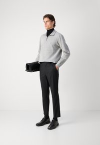 Šviesiai pilka bliuzono su užtrauktuku viršuje, juodos kelnės ir juodi batai. Modelis laiko juodą rankinę; elegantiškas dizainas, minkšta tekstūra ir pritaikytas kirpimas.
