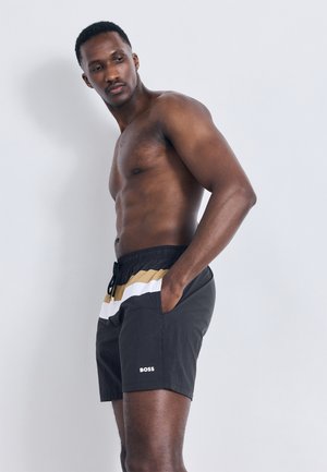 Homme torse nu debout devant un fond gris clair uni, portant un short de bain BOSS noir, blanc et beige, les mains dans les poches.