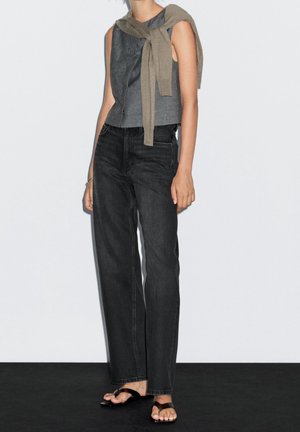 Mannequin portant un top gris sans manches, un jean taille haute foncé, des sandales noires, avec un pull beige posé négligemment sur les épaules.