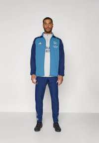 Kék és tengerészkék sportdzseki fehér részletekkel és logóval, hozzá illő nadrággal, oldalcsíkkal és fekete cipővel.