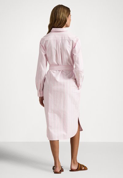 Sieviete, tērpusies rozā un baltā svītrainā krekla kleitā ar jostu, garām piedurknēm un sānu šķēlumu, valkājot brūnus atvērtā tipa sandales, stāvot ar muguru pret skatītāju.