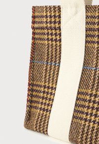 Τσάντα tote με καφέ, κίτρινο και κόκκινο καρό ύφασμα και φαρδιά λευκή υφασμάτινη αναδιπλούμενη strap σε λευκό φόντο.