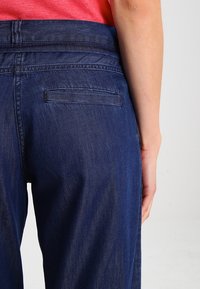 Tmavě modré džíny s rovnými nohavicemi, hladkou texturou a jednou zadní kapsou. Pas je mírně elastický.