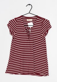 Camisa de manga corta a rayas burdeos y crema con escote en forma de V y detalle de cordones en el frente. Hecha de tela acanalada, colgando de un gancho negro.