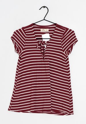 Burgundowa i kremowa w paski koszula z krótkim rękawem, z dekoltem w serek i detalem sznurowania z przodu. Wykonana z prążkowanej tkaniny, wisząca na czarnym wieszaku.