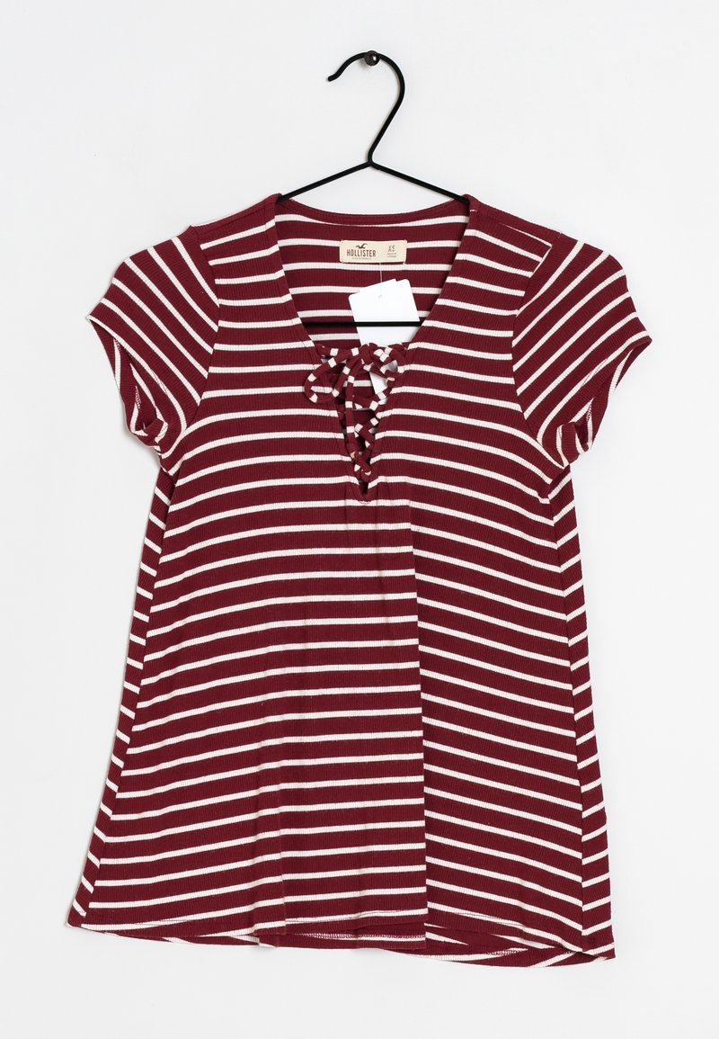 Camisa de manga corta a rayas burdeos y crema con escote en forma de V y detalle de cordones en el frente. Hecha de tela acanalada, colgando de un gancho negro.