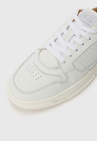 Zapatilla blanca de cuero con puntera perforada, cordones blancos planos y suela beige. Presenta un acento rectangular en el lateral y una marca en la lengua.
