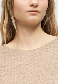 Gros plan sur le bas du visage, le cou et les épaules d'une femme portant un haut texturé beige avec de longs cheveux blonds.