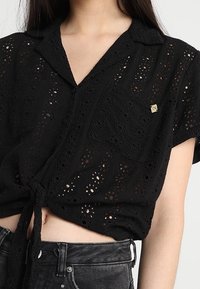 Camicia nera a maniche corte in tessuto ad occhio di pernice, con colletto con bottoni, tasca frontale e dettaglio di nodo sul davanti.