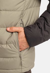 Veste matelassée beige légère avec une manche sombre. Comprend une poche zippée et une texture matelassée pour chaleur et isolation.