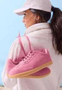 Pink suede sneakers with a gum rubber sole, featuring round laces and subtle branding, held by a person wearing a light hoodie.