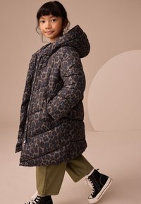 Kurtka puchowa w ciemnym niebieskim kolorze z wzorem w panterkę, wyposażona w kaptur, zapięcie na zamek błyskawiczny oraz pikowany design. Połączona z zielonymi spodniami i czarnymi trampkami.