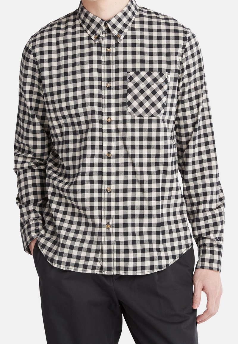 Chemise à carreaux noir et blanc avec col, manches longues et poche poitrine. Comprend des boutons marron et une texture douce.