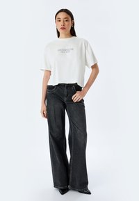 Camiseta corta blanca con la impresión "BROOKLYN New York", combinada con unos jeans de tiro alto de pierna ancha negros y tacones negros de punta.