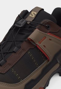 Κοντινό πλάνο καφέ και μαύρου πεζοπορικού παπουτσιού με κορδόνια ταχείας έλξης, ενισχυμένη μύτη και επένδυση στον αστράγαλο σε λευκό φόντο.