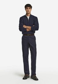 Navy-Blue Zip-Up-Pullover, kombiniert mit navyfarbenen Hosen, getragen mit braunen Lederschuhen mit hellbraunen Schnürsenkeln. Das Model steht mit verschränkten Armen.