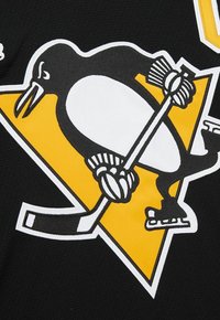 Pinguin in Schlittschuhen und Handschuhen, der einen Hockeyschläger hält, vor einem gelben Dreieckshintergrund auf schwarzem Stoff.