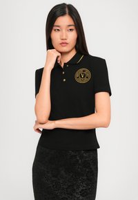 Young woman with straight black hair wearing a black polo shirt with gold Versace Jeans Couture logo and a black floral patterned skirt.