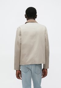 Ανοιχτό μπεζ μπουφάν με δομημένο σχέδιο, που διαθέτει καφέ γιακά από κορντόρι, μακριά μανίκια και ίσια γραμμή, συνδυασμένο με ανοιχτό μπλε τζιν.