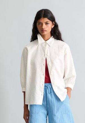 Kobieta o długich ciemnych włosach, ubrana w luźną białą koszulę zapinaną na guziki, nałożoną na czerwoną bluzkę i jasnoniebieskie spodnie w paski, stojąca na jednolitym tle.