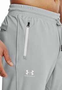 Svetle šedé sportovní kalhoty s texturovaným povrchem, s boční kapsou na zip, elastickým pasem a bílým logem Under Armour.