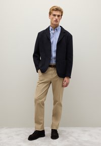 Uomo in giacca blu marina, camicia a righe blu con bottoni, pantaloni beige e scarpe marrone scuro in piedi contro un muro chiaro e semplice.