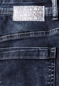 Donkerblauwe denim jeans met een vervaagde textuur. Voorzien van een wit etiket met patroontekst en een contrasterende stikseldetail.