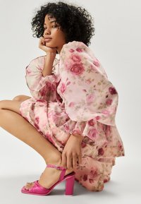 Vestido floral rosa con mangas abullonadas y volantes en capas; combinado con sandalias de tacón rosa brillantes con tira en el tobillo y detalle de nudo.