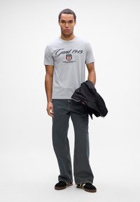 T-shirt de algodão cinza com o logótipo e gráfico "Gant 1949" em preto, combinado com jeans escuros largos e sapatilhas pretas.