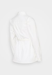 Vit långärmad skjorta med knäppkrage, baktillveck och ett bälte i midjan, med en mjuk bomullstextur.